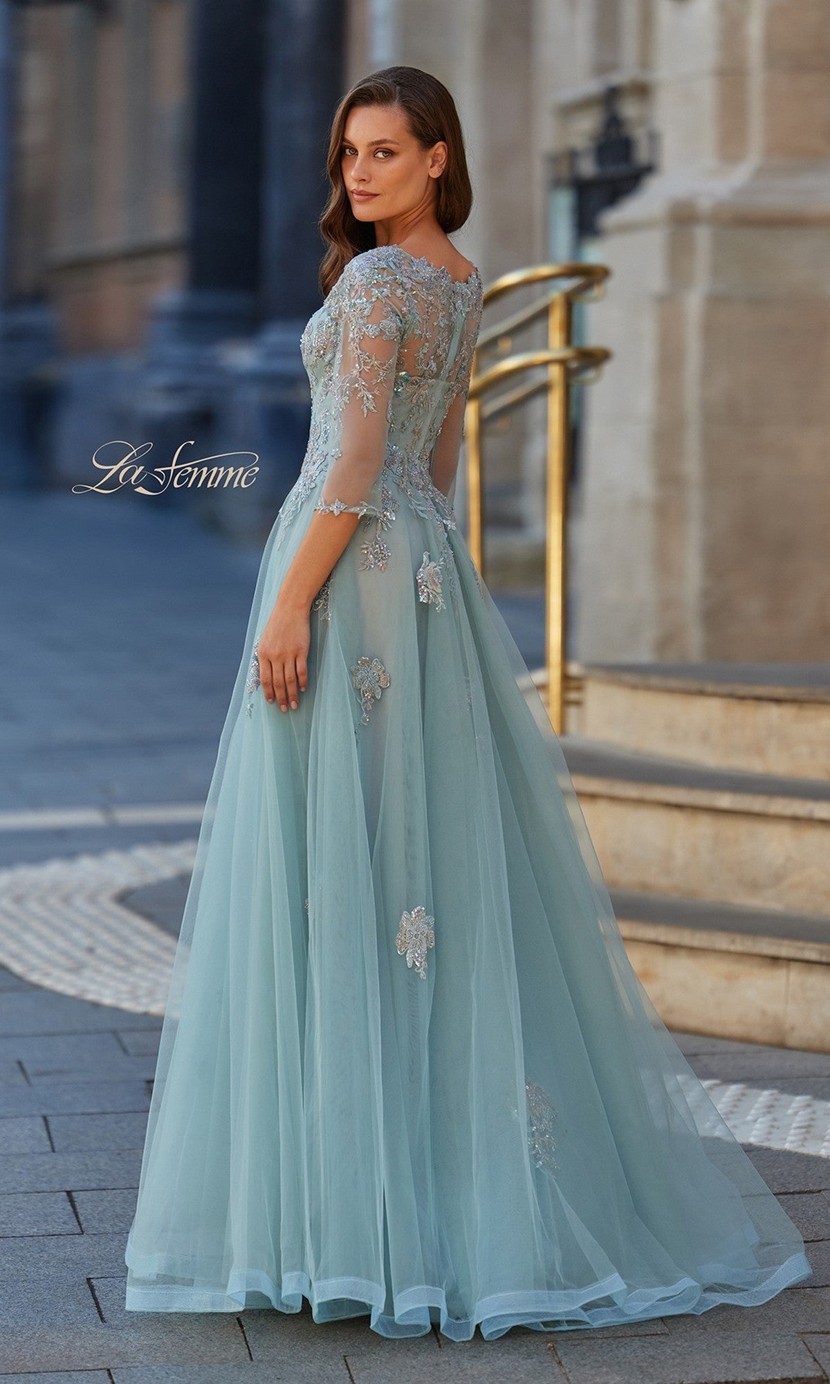 A woman stands outdoors on a paved path, looking over her shoulder in the La Femme 33536 long formal dress—light blue, floor-length tulle with floral embroidery.